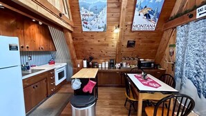 Interior d'una cabana amb parets de fusta, zona de cuina amb nevera, microones i rentamics, taula de dinar amb caires i pòsters de Whistler i Montana.