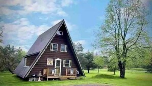 Una casa de fusta de forma triangular amb un porxo, envoltada d'arbres i un cel blau.
