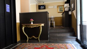 A hotel lobby with a black reception desk, a decorative table with a vase, a patterned rug, a staircase, and a television on the wall. - Hotel Valentino Du Parc (Turin)