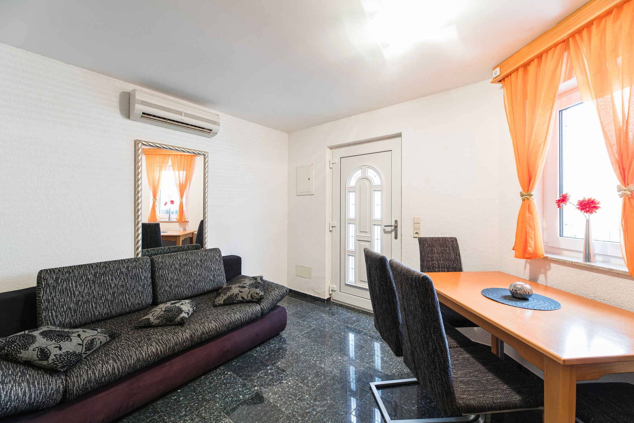 Een kamer met een bank, een eettafel en stoelen. Er is een deur en een raam met gordijnen.