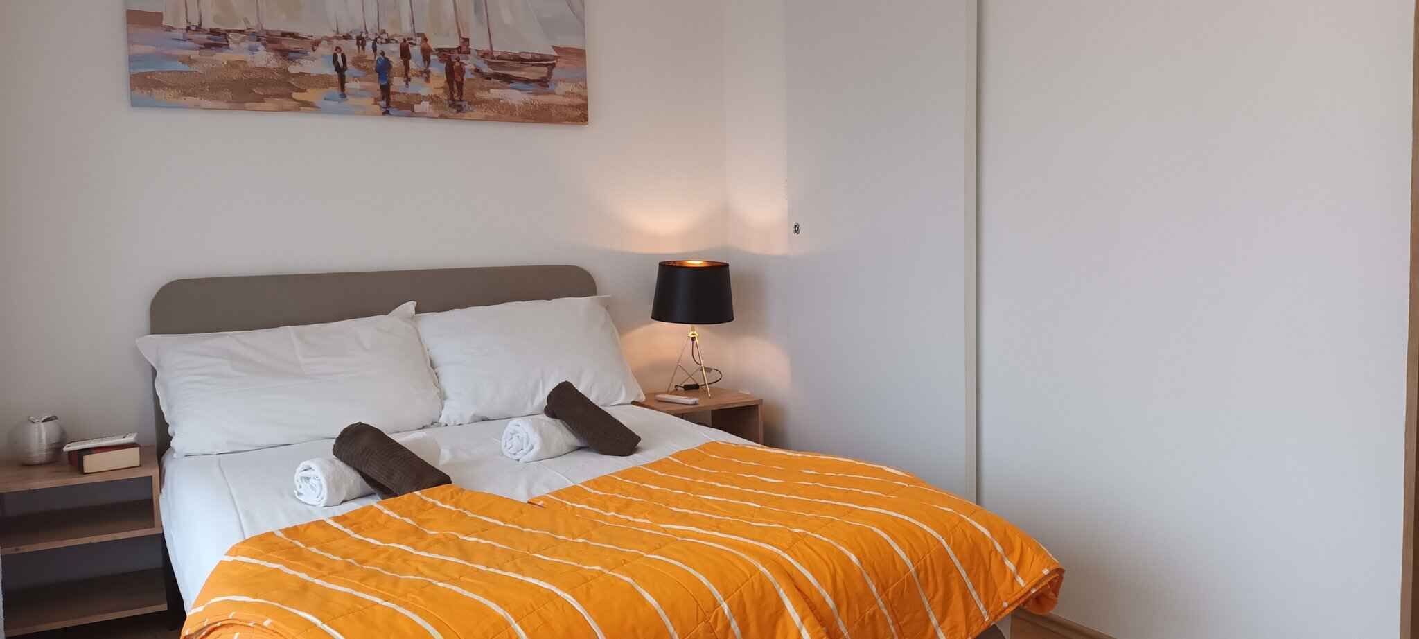 A neatly made bed with white and brown pillows, an orange striped blanket, and a painting of a beach scene on the wall.