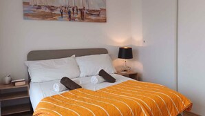 Ein ordentlich bezogenes Bett mit weißen und braunen Kissen, einer orangefarbenen Streifendecke und einem Strandbild an der Wand.