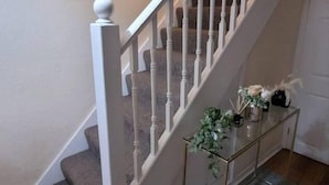 Een trap met witte balustrades, een klein console tafeltje met een plant, en twee bloemige kunstwerken aan de muur.