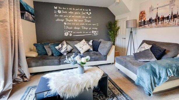 Un soggiorno moderno con un divano grigio, un tavolino da caffè e un pouf bianco in similpelle, e una parete decorata con citazioni motivazionali.