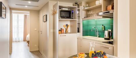 Cocina compacta con microondas, pileta y salpicadero verde. Hay un comedor con una mesa dispuesta para el desayuno, incluyendo frutas y un periódico.