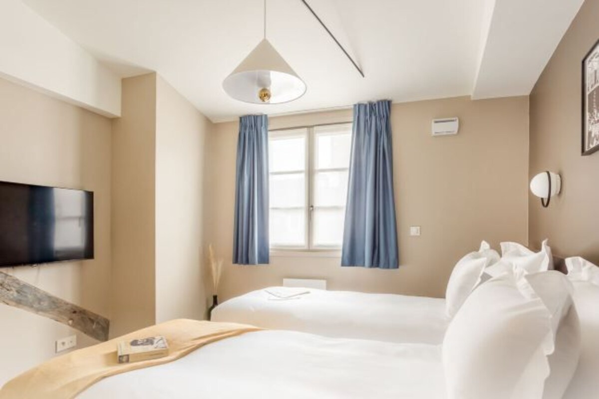 A hotel room with two beds, a flat-screen TV mounted on the wall, a window with blue curtains, and a ceiling light fixture.