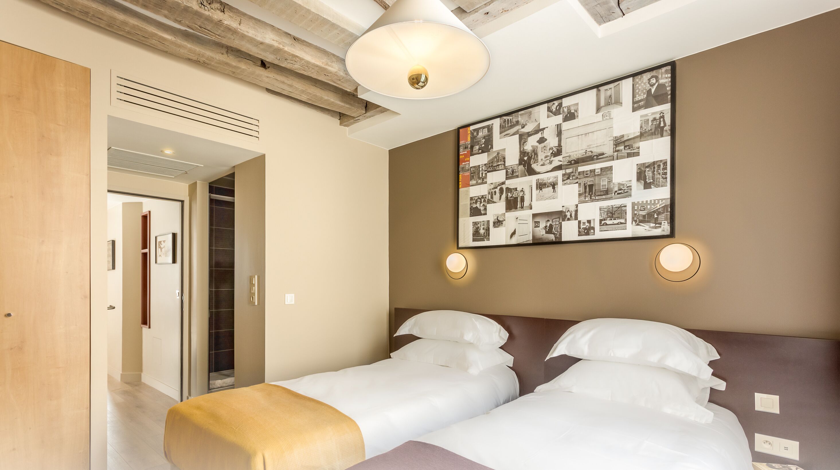 Une chambre d’hôtel avec deux lits, un plafond en bois, un collage de photos accroché au mur et une porte donnant sur une autre pièce.