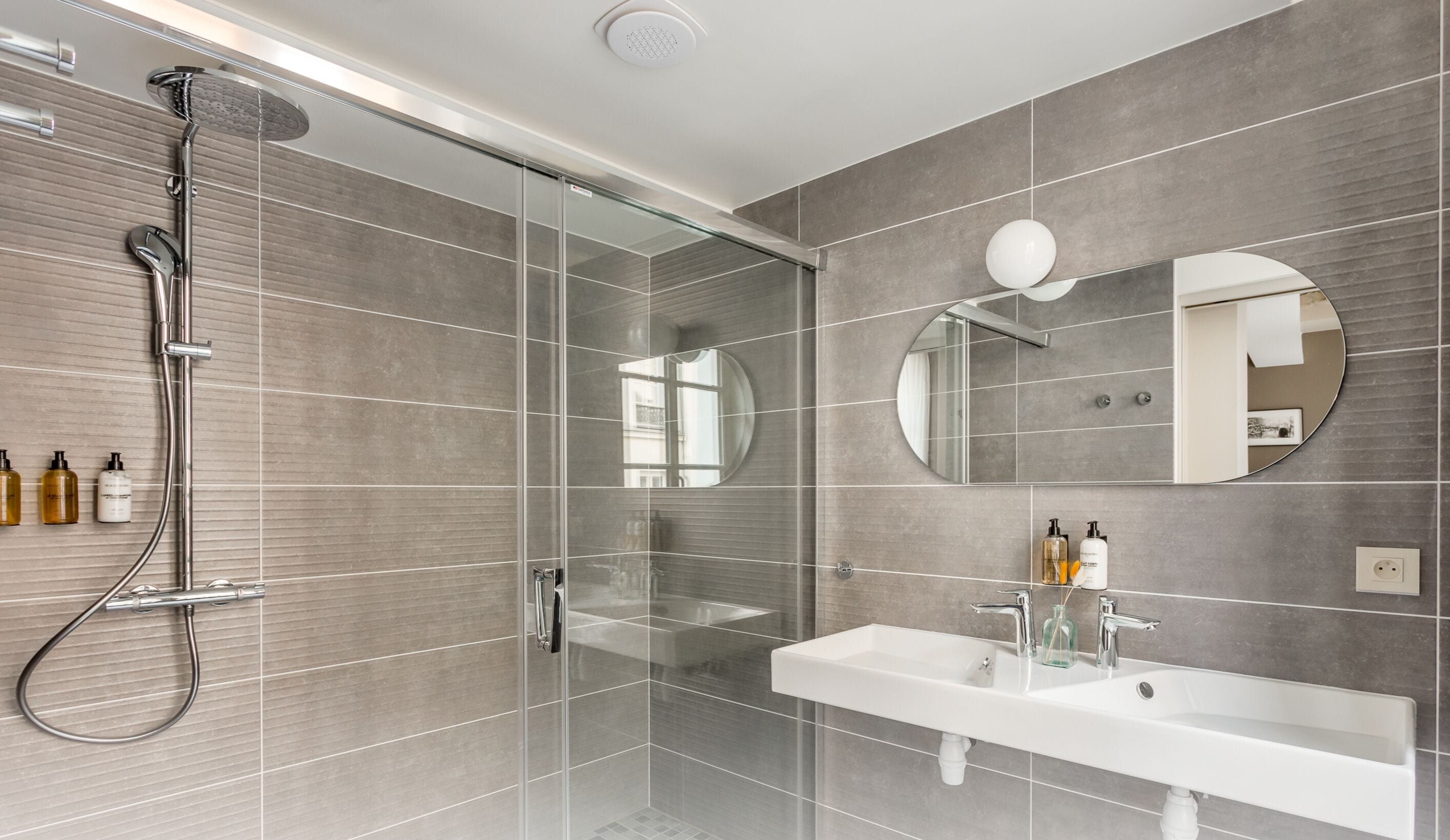 Une salle de bain moderne avec une douche à parois de verre, un grand miroir et un lavabo blanc sur lequel sont posés deux flacons de shampoing.