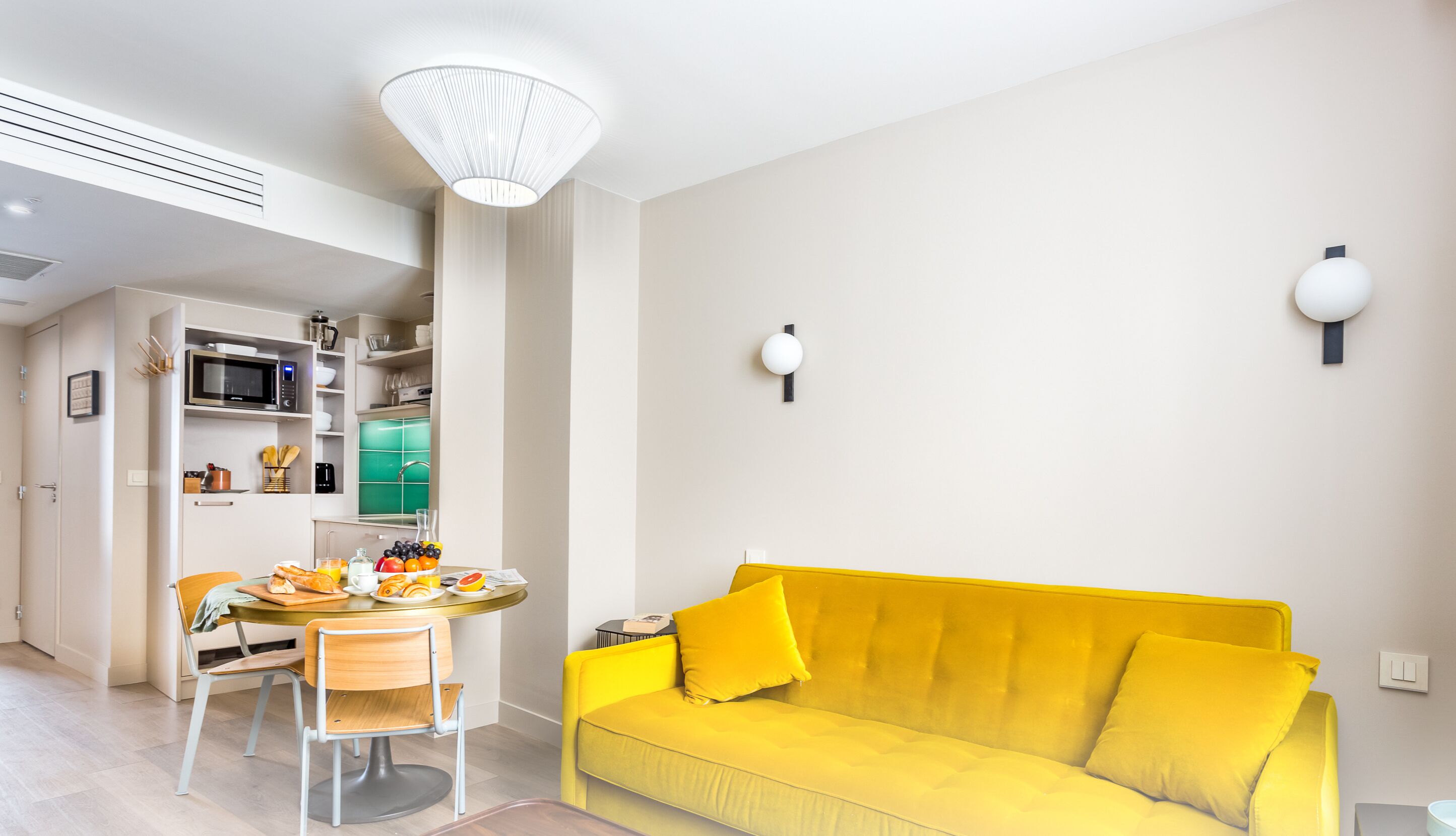 A bright yellow sofa, a dining table with chairs, a kitchen area with a microwave and shelves, and a white ceiling light.