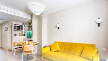 A bright yellow sofa, a dining table with chairs, a kitchen area with a microwave and shelves, and a white ceiling light.
