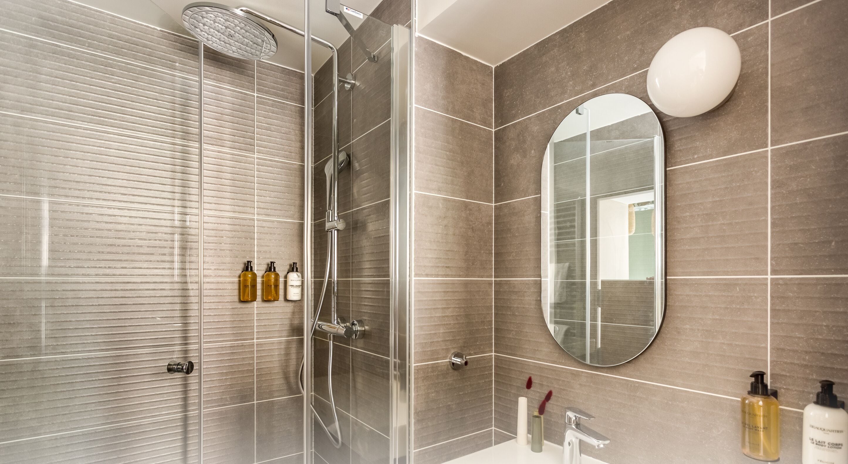 Une salle de bain moderne avec une douche à parois de verre, un grand miroir et des articles de toilette posés sur le meuble-lavabo.