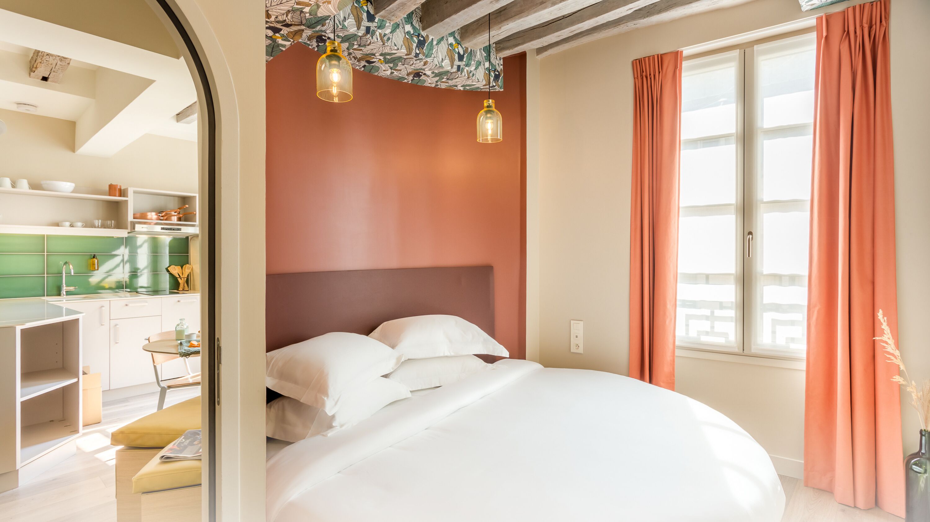 A bedroom with a large bed, a window with curtains, and a view of the kitchen.