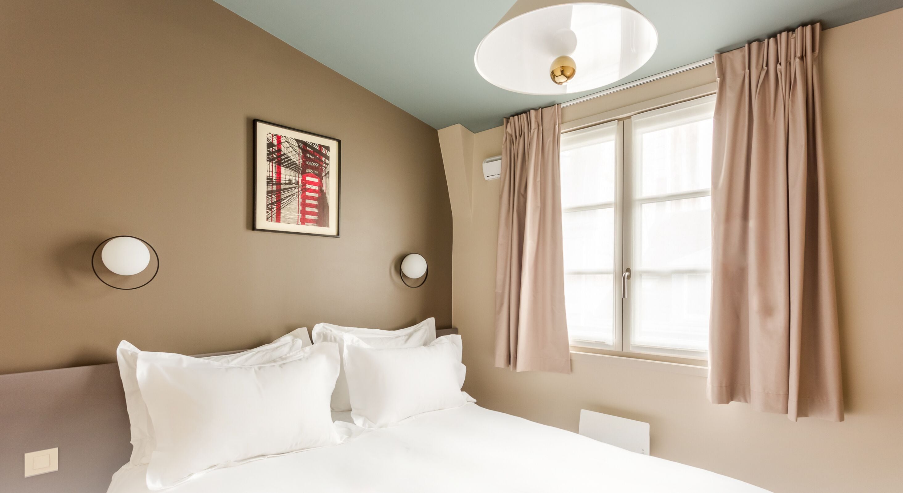 Une chambre d’hôtel comprenant un lit, deux appliques murales, un luminaire au plafond, une fenêtre avec des rideaux et un climatiseur fixé au mur.