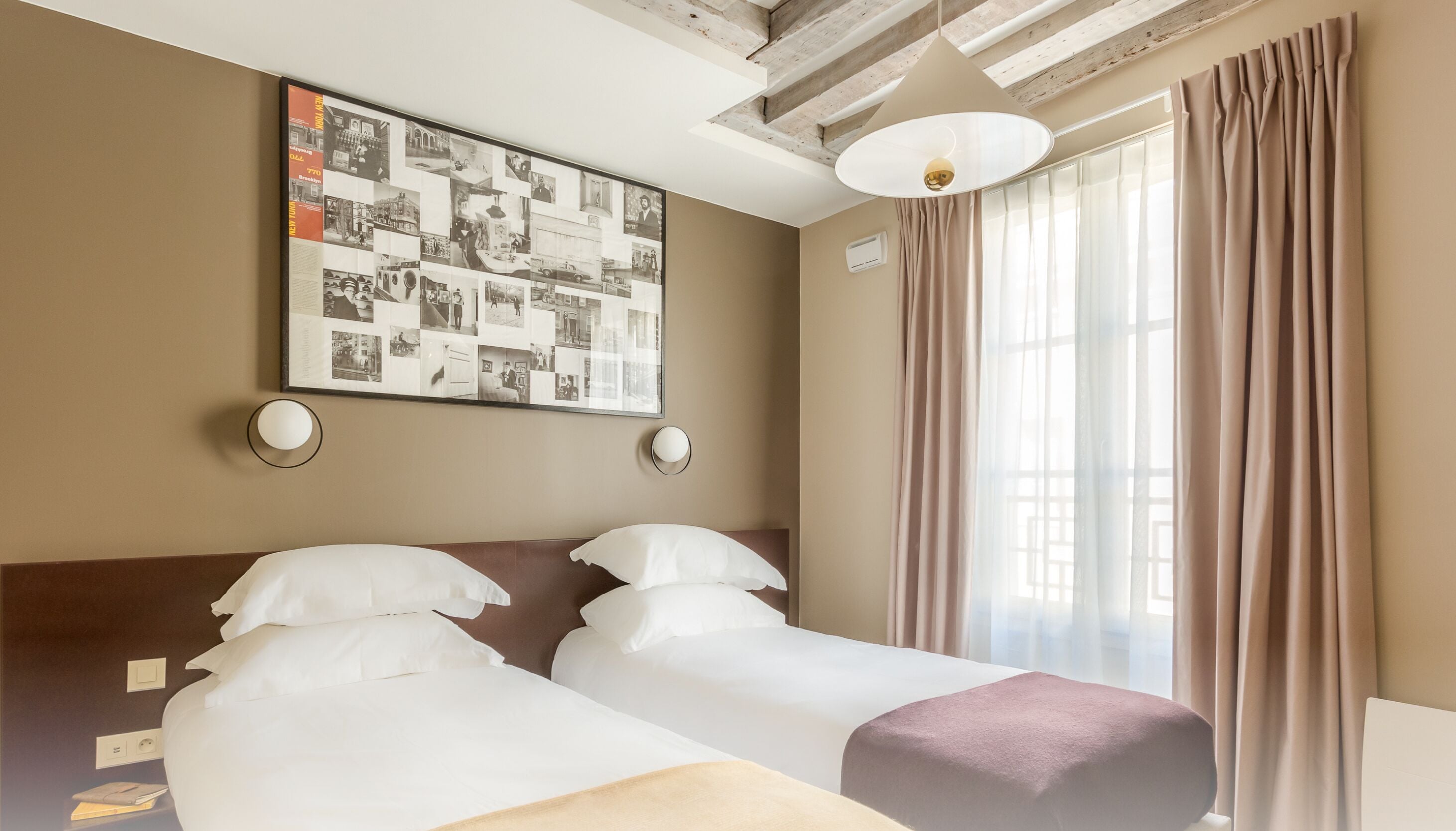 A hotel room with two beds, a large collage artwork on the wall, a window with curtains, and a ceiling with exposed beams.