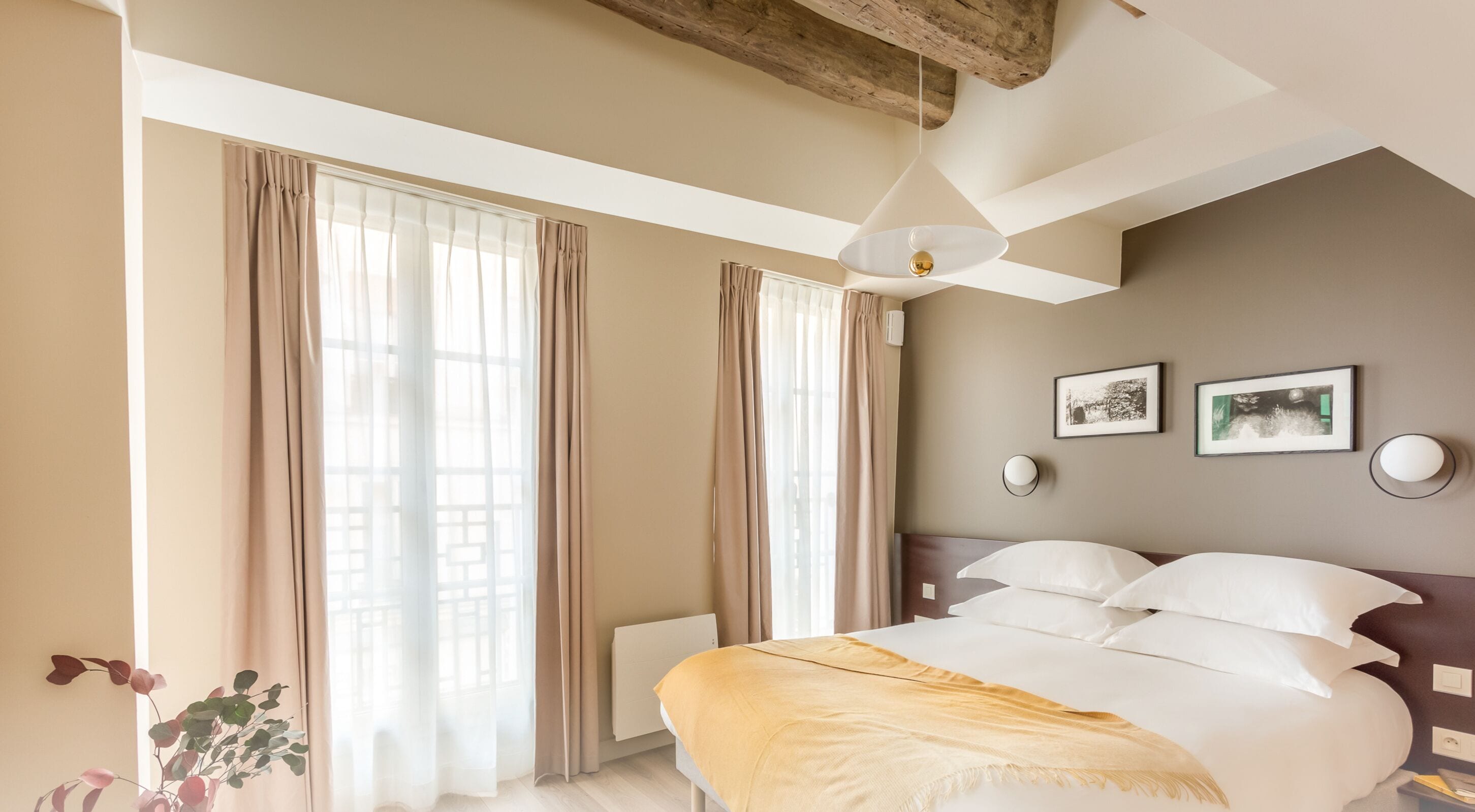 Habitación de hotel con cama, dos ventanas con cortinas, un radiador, una lámpara colgante y dos cuadros en la pared.