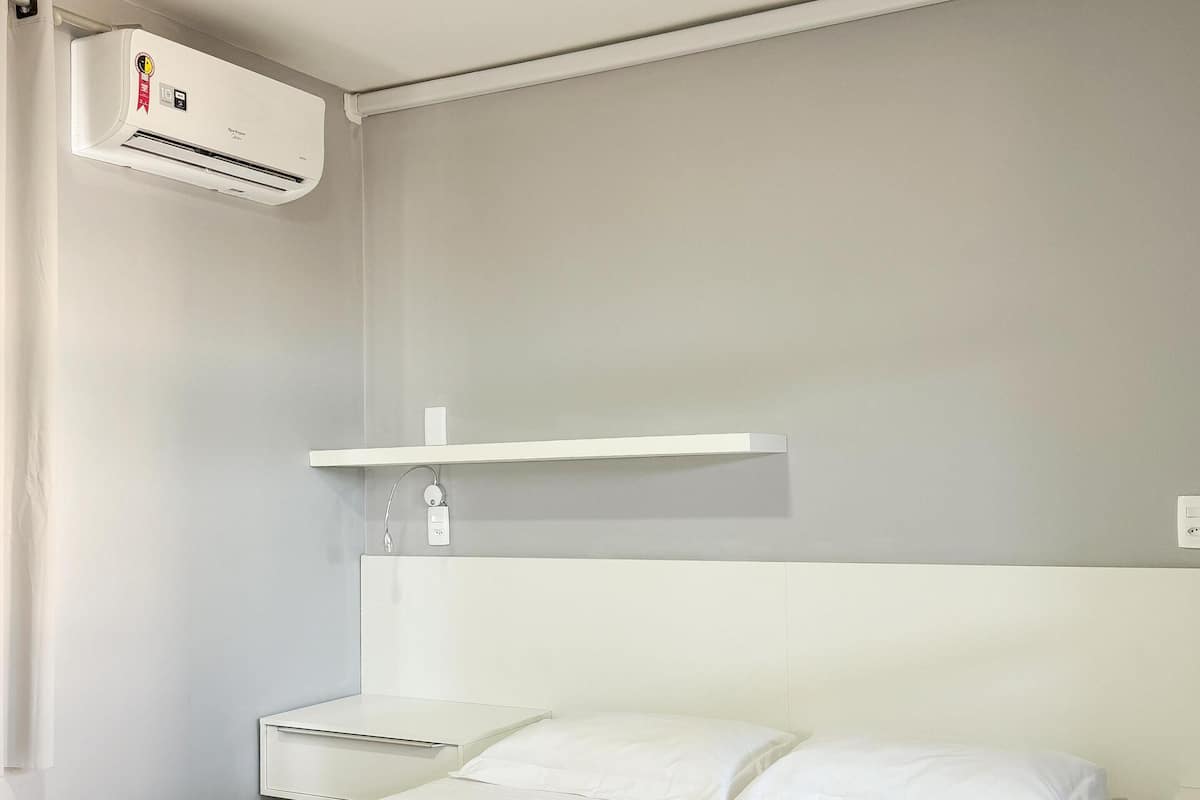 A hotel room with a bed, a bedside table, a wall-mounted air conditioner, and a shelf with a small item on it.