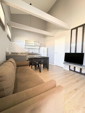 Una sala moderna con un sofá, una mesa de comedor con sillas, una zona de cocina con microondas y refrigerador, y un televisor sobre un mueble.