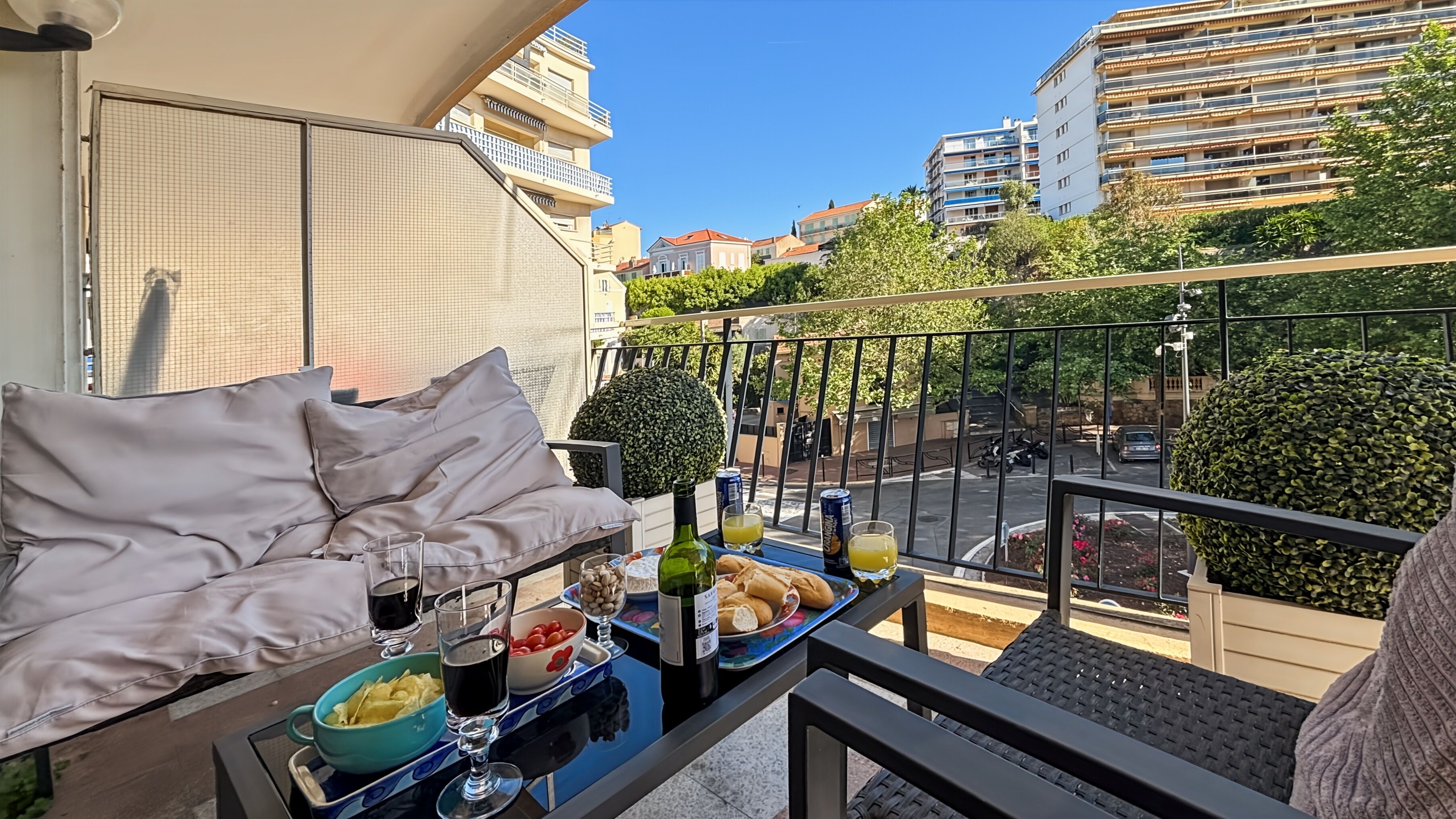 A balcony with a sofa, a table with food and drinks, and a view of buildings and greenery.