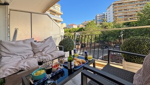 A balcony with a sofa, a table with food and drinks, and a view of buildings and greenery. - Comfortable and fully equiped two bedroom apartment (Cannes)