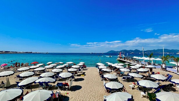 A beach with rows of umbrellas and chairs, a pier extending into the sea, and boats in the background. - Comfortable and fully equiped two bedroom apartment (Cannes)