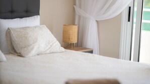 A neatly made bed with a headboard, a bedside table with a woven lamp, and a view of the outdoors through a window with white curtains. - Narcissos Nissi Beach Apartment E9 (Ayia Napa)