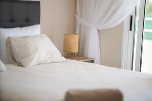 Ein ordentlich bezogenes Bett mit Kopfteil, ein Nachttisch mit Lampenschirm aus Weidengeflecht und Blick auf eine grüne Außenanlage durch ein Fenster mit weißen Vorhängen.