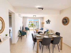 A modern dining area with a large table set for six, a sofa, and a balcony with a view of buildings. - Narcissos Nissi Beach Apartment E9 (Ayia Napa)
