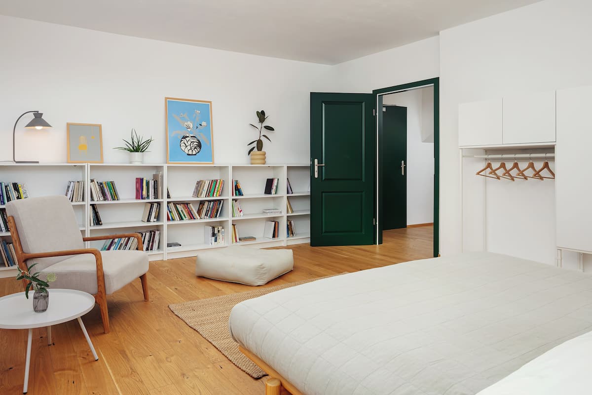A bedroom with a bed, a chair, a small table, a bookshelf, and a green door.