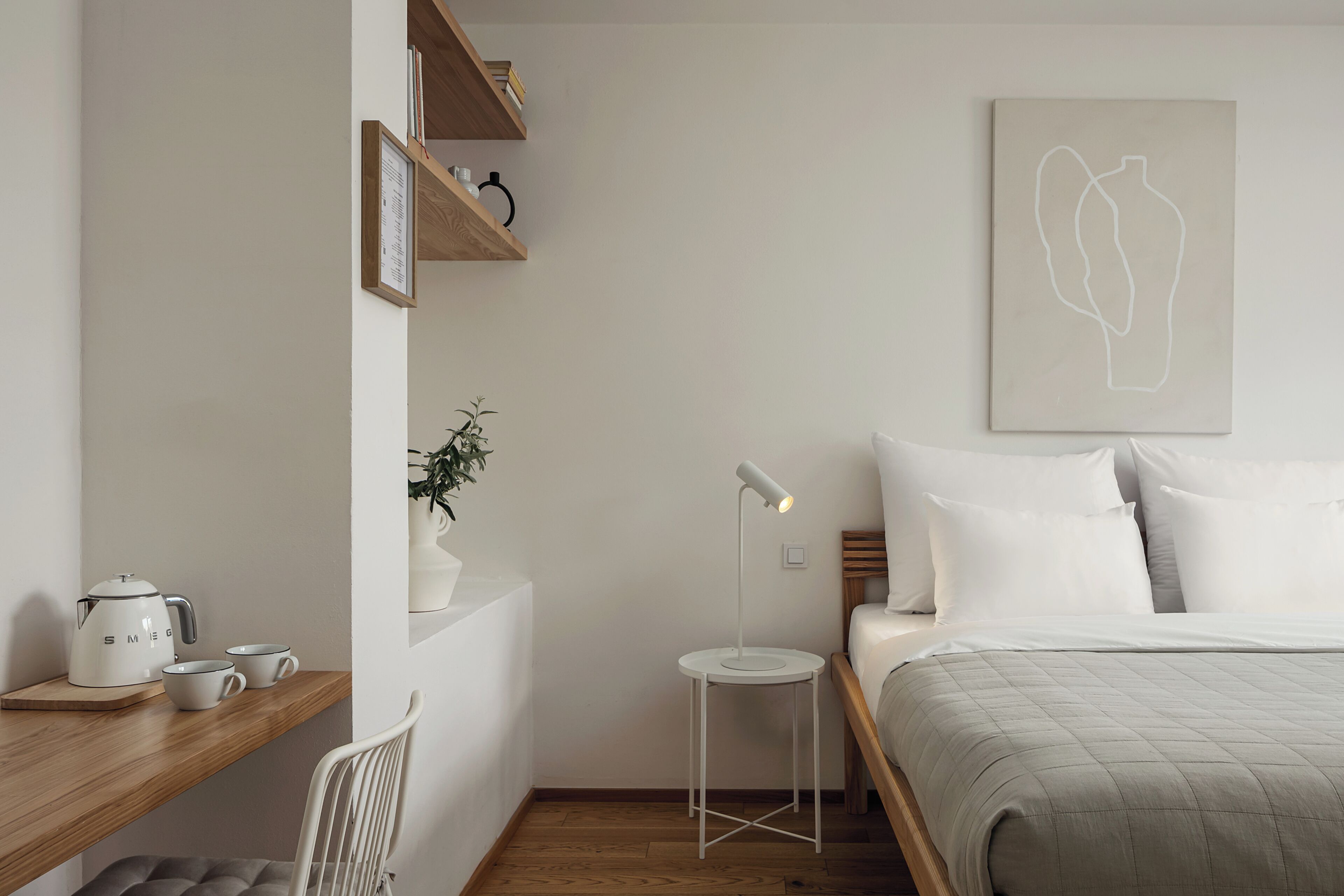 A minimalist bedroom with a bed, bedside table, lamp, and a wall-mounted shelf with books and a plant.