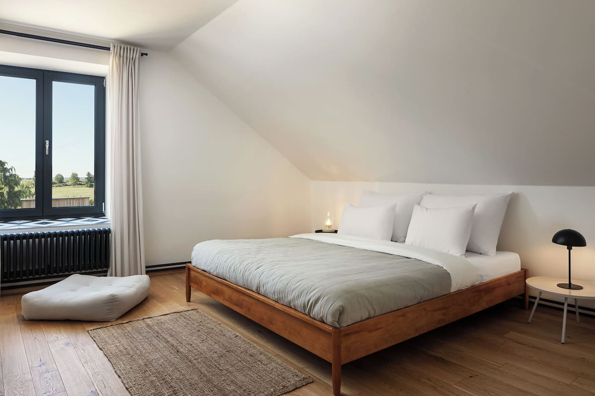A bedroom with a wooden bed, a nightstand, a lamp, a window with curtains, and a view of the outdoors.
