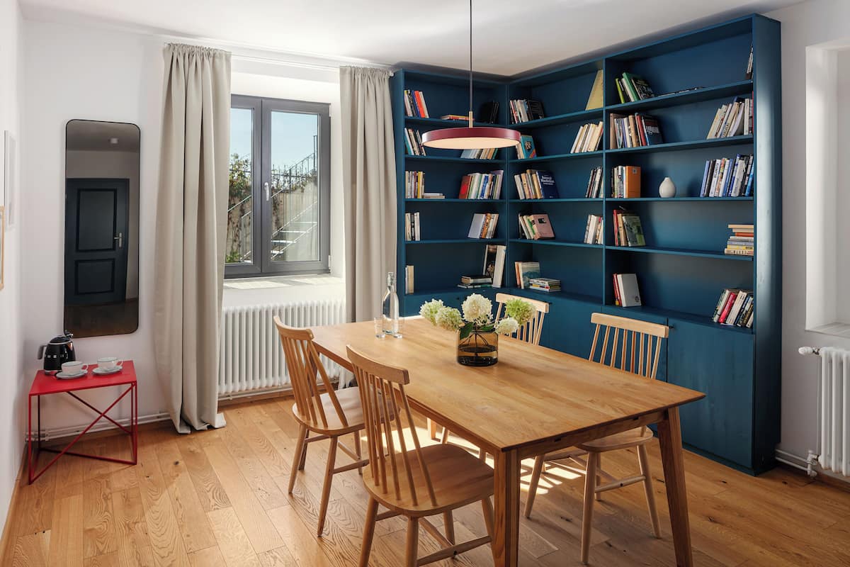 A dining area with a wooden table, chairs, a red side table, a blue bookshelf, and a window with curtains.
