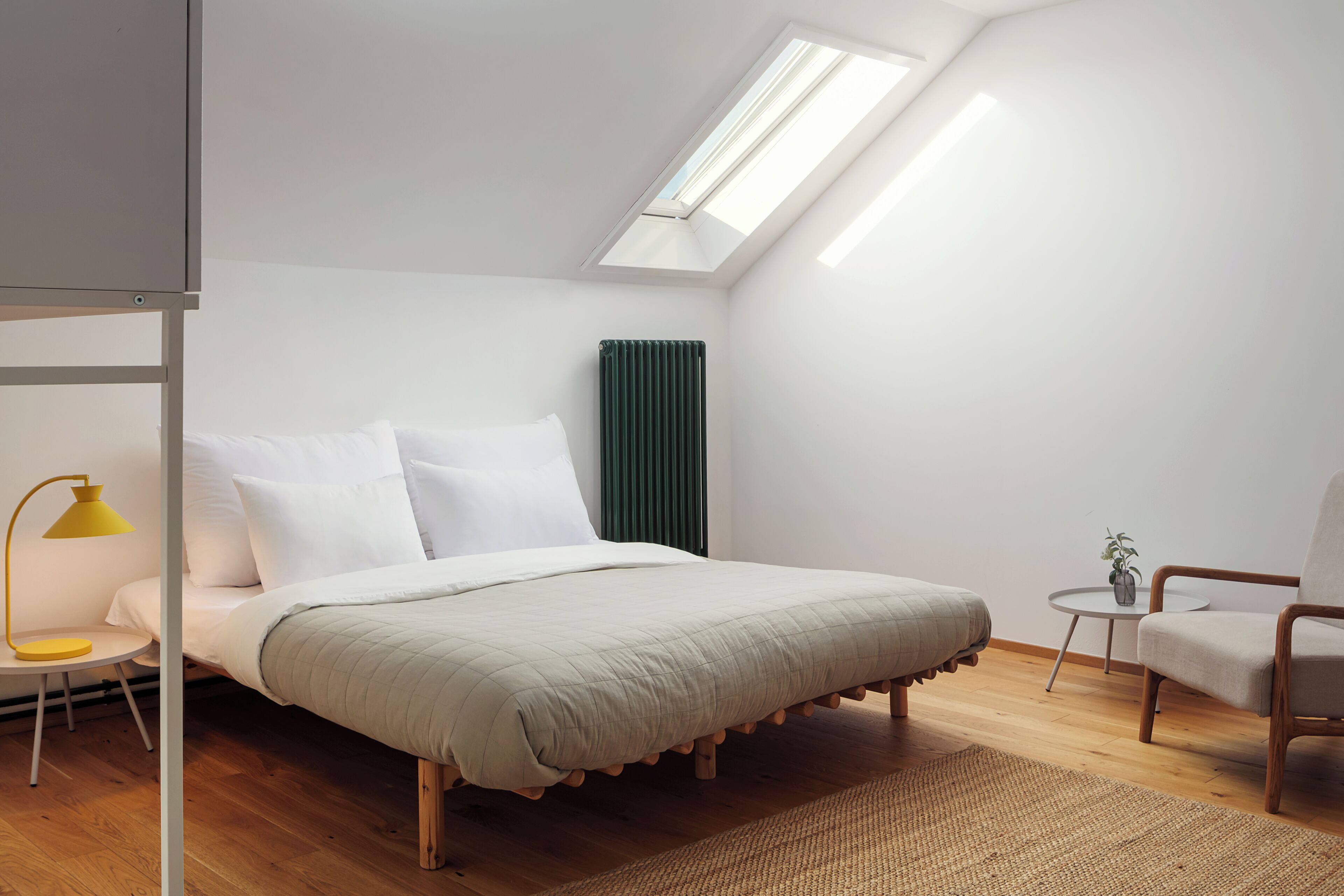 Une chambre à coucher comprenant un lit, une chaise, une petite table, une lampe, un radiateur et une lucarne.