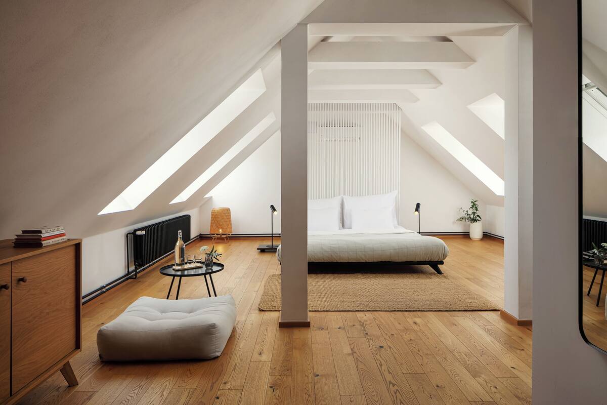 A modern loft bedroom with a sloped ceiling, a bed, a small seating area, and a radiator.
