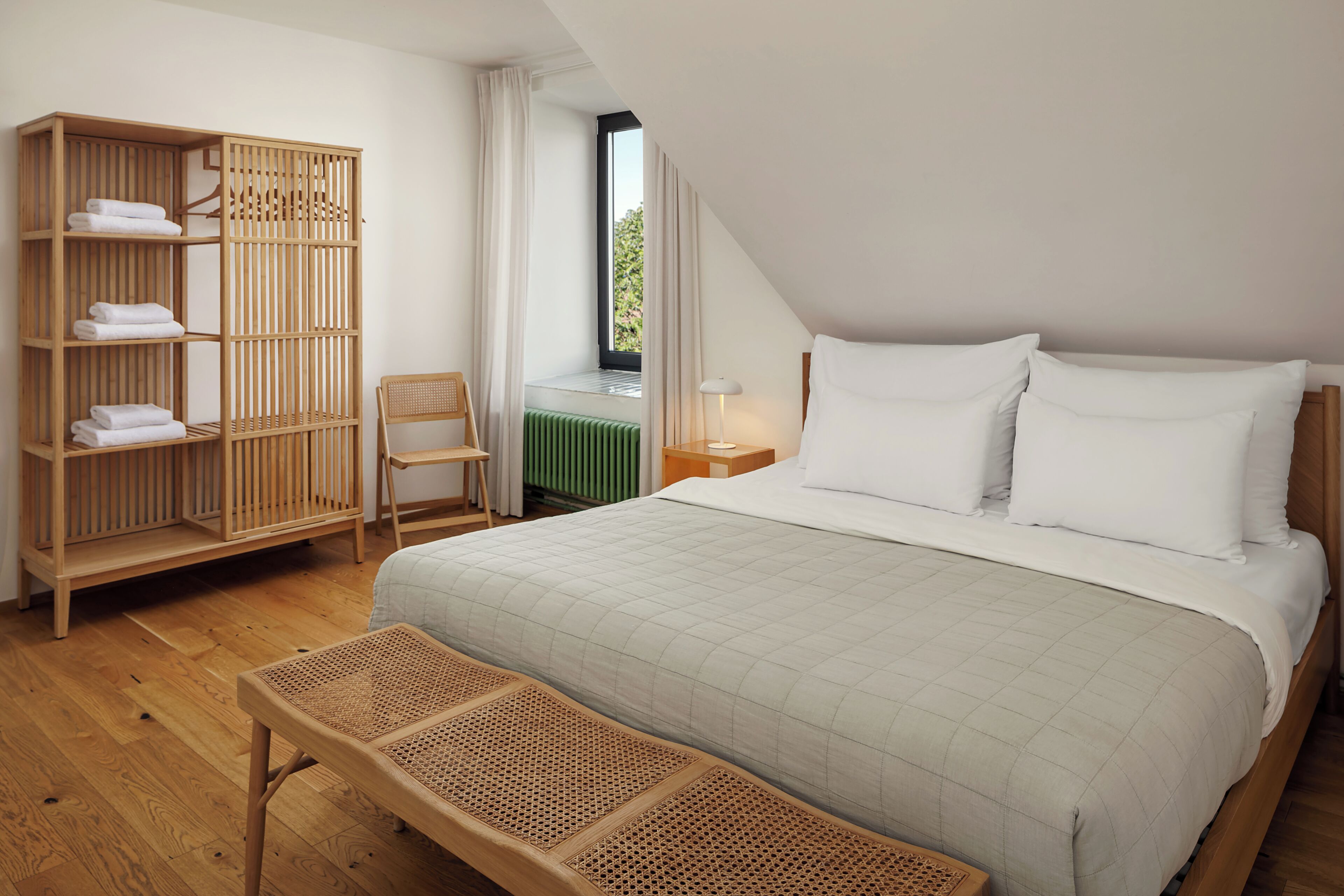 A bedroom with a bed, a wooden wardrobe, a bench, a chair, and a window with curtains.