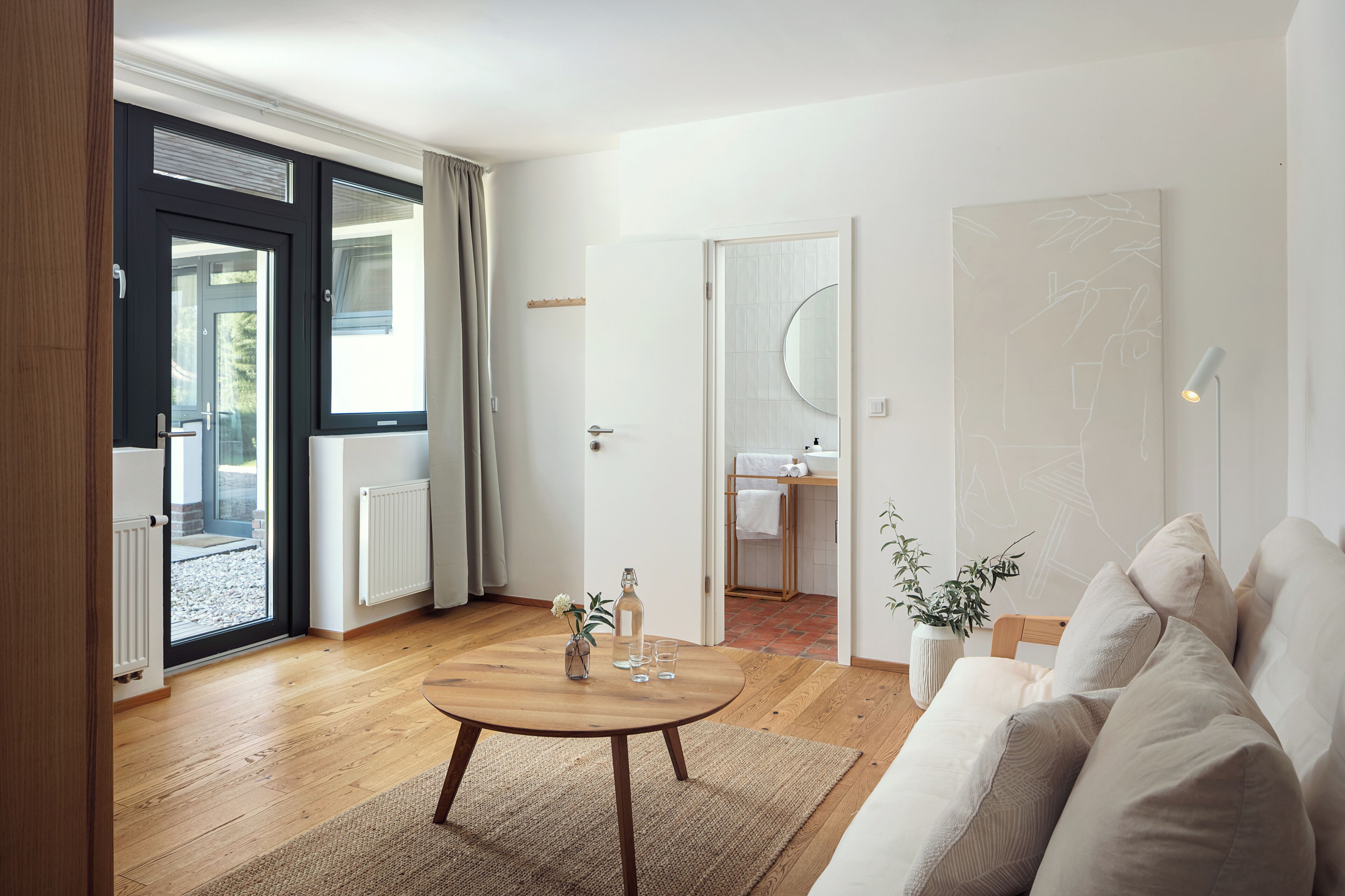 A modern living room with a wooden floor, a round coffee table, a sofa, a sliding glass door, and a bathroom visible through an open door.
