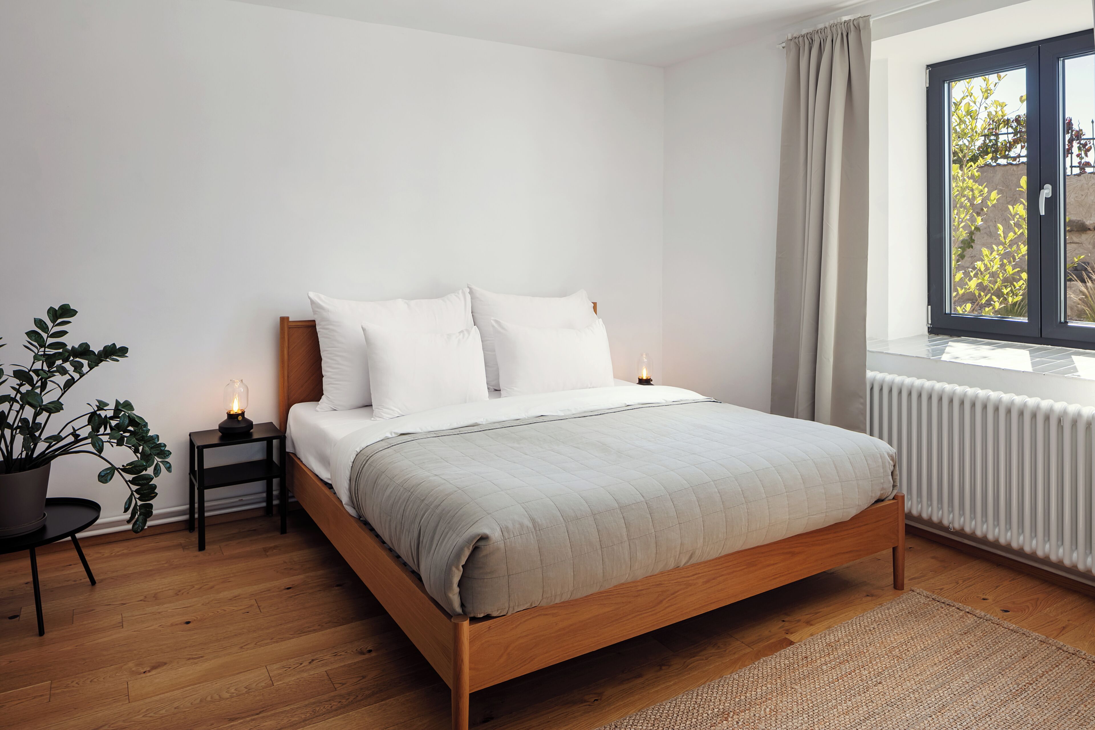 Um quarto com cama de madeira, roupa de cama branca, uma pequena mesa de cabeceira com luminária, uma planta em vaso e uma janela com cortinas.