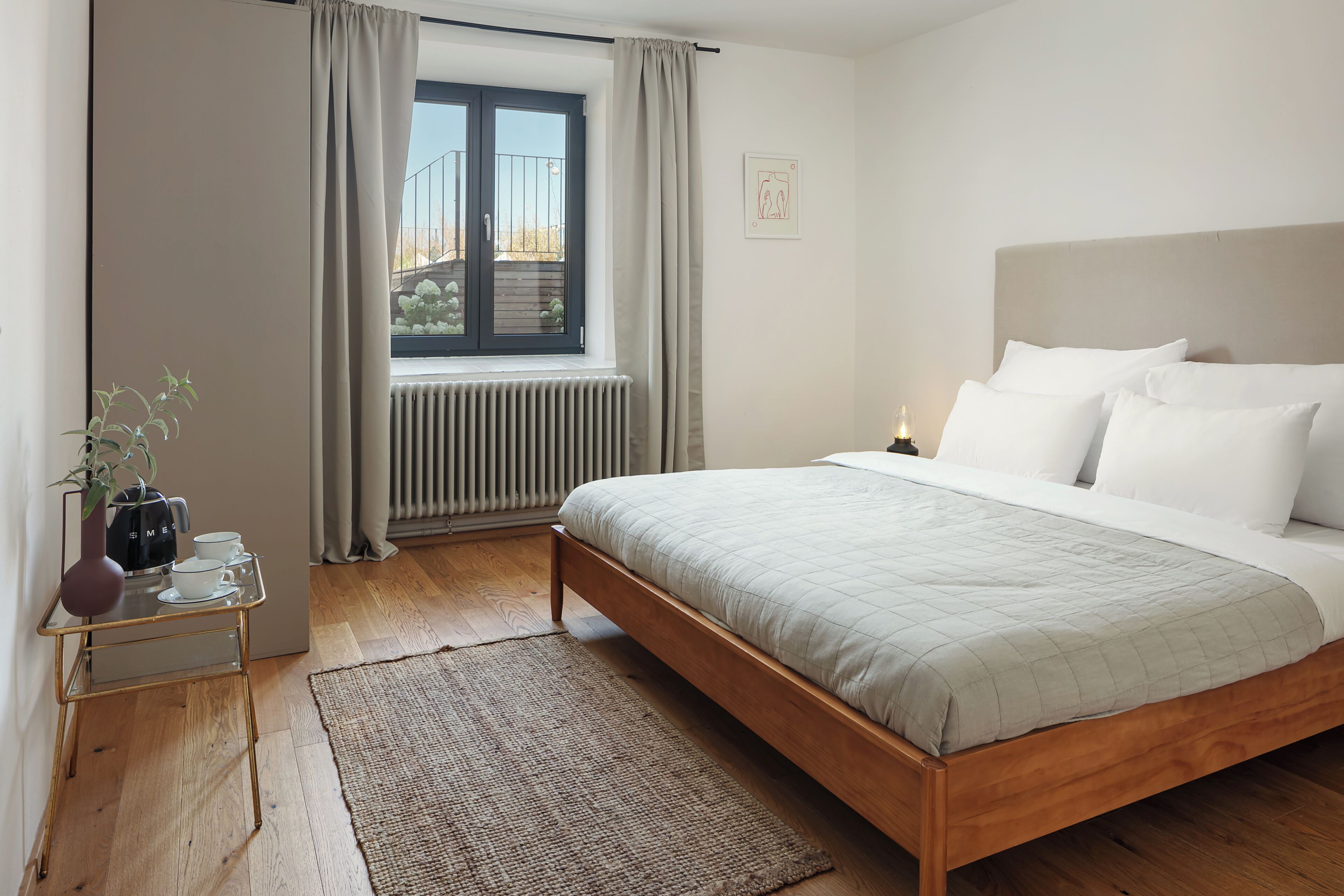 Une chambre à coucher comprenant un lit, un radiateur, une fenêtre avec des rideaux, une table de chevet avec une plante et un lampadaire fixé au mur.