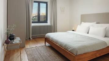 Une chambre à coucher comprenant un lit, un radiateur, une fenêtre avec des rideaux, une table de chevet avec une plante et un lampadaire fixé au mur.