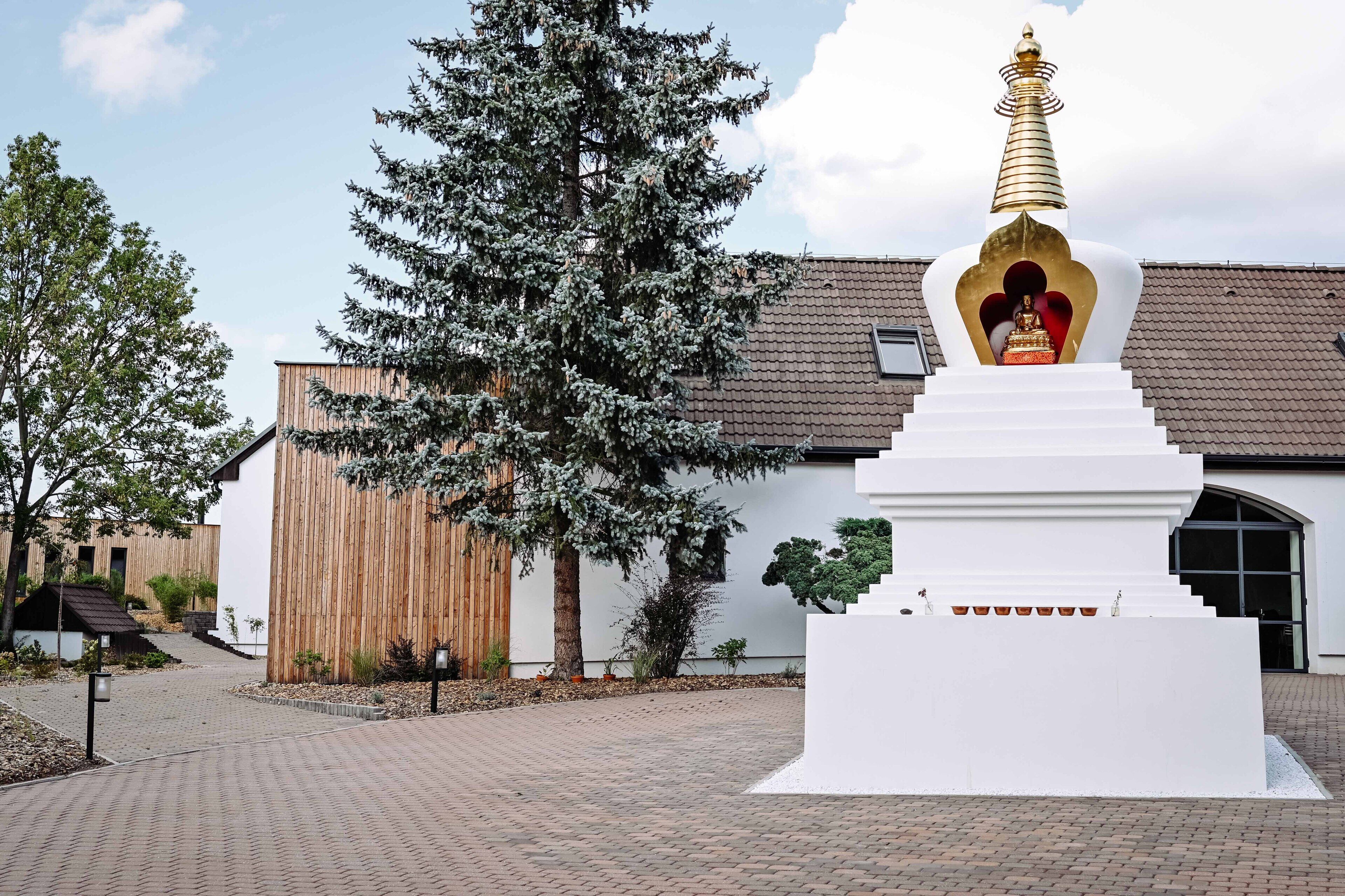 Un stupa blanc surmonté d’une coupole dorée, un bâtiment en bois et un grand arbre à l’avant.