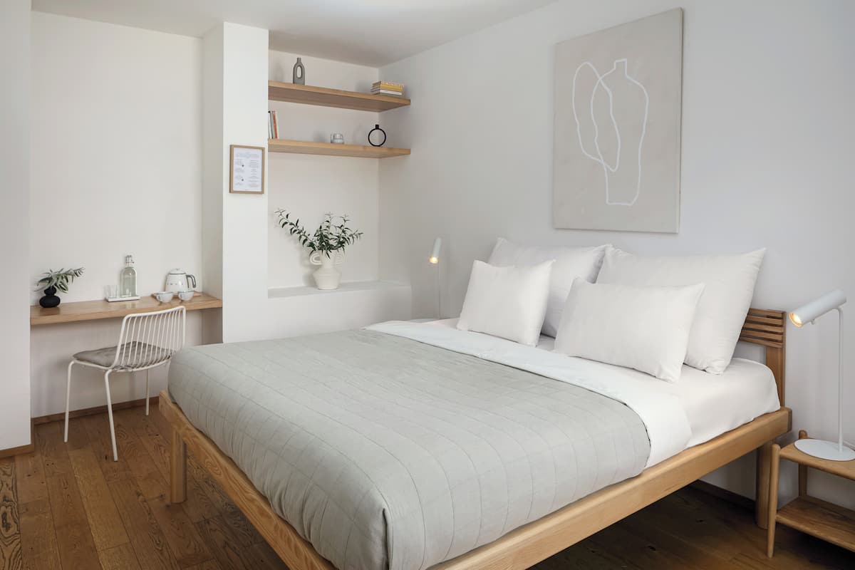A bedroom with a bed, a desk, a chair, a bedside table, a lamp, and shelves with books and decorative items.