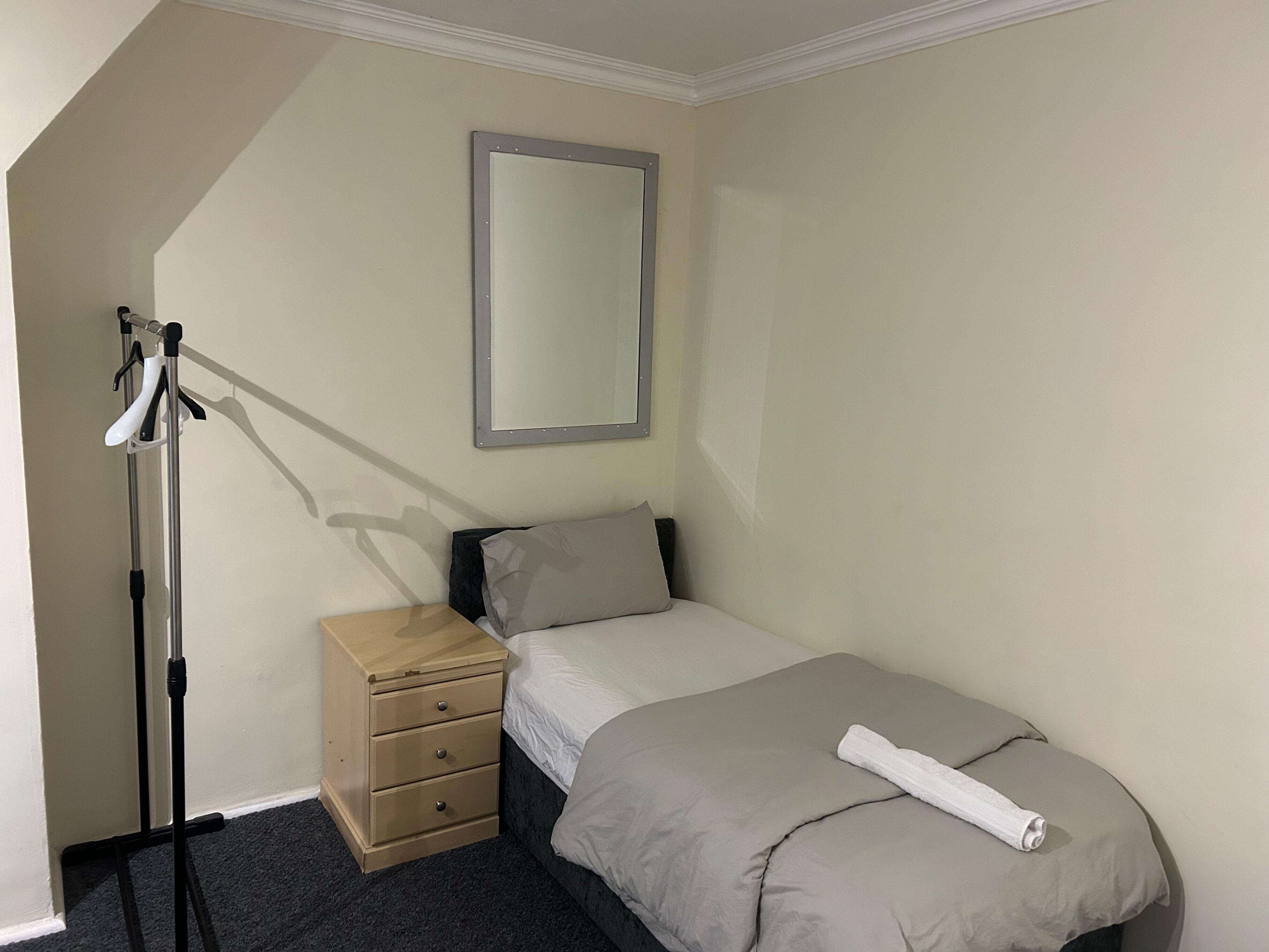 A small, neatly arranged bedroom with a single bed, a bedside table, a mirror, and a coat rack.