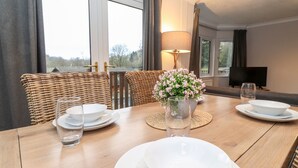 Una mesa de comedor dispuesta con platos, tazones y vasos blancos, un florero con flores, sillas de mimbre y un televisor al fondo.