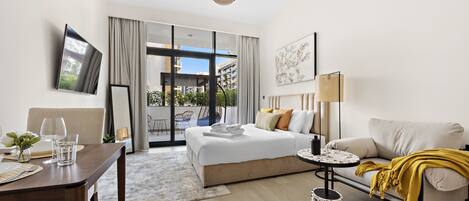 A modern hotel room with a bed, sofa, dining table, and a TV mounted on the wall.