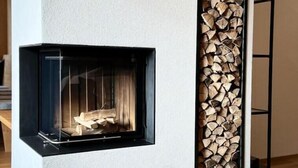 Ein moderner Kamin mit Glasfront, in eine weiße Wand integriert, mit einem Holzstapel daneben.