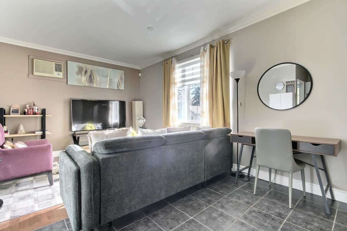 Un salon avec un canapé gris, un téléviseur, un bureau avec une chaise, un grand miroir et une fenêtre avec des rideaux.