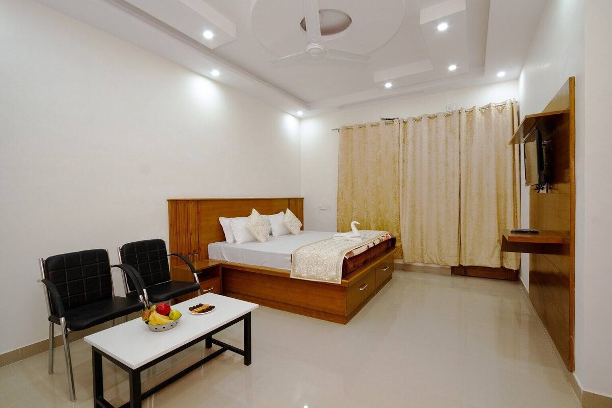 A hotel room with a bed, two chairs, a small table with fruit, and a TV.