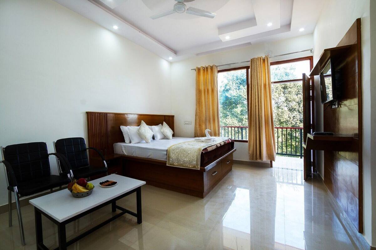 A hotel room with a bed, a desk with a fruit bowl, a chair, a TV, and a balcony with curtains.