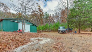 A rural setting with a green metal shed, a dirt path, and a vehicle parked near trees. - Bunkhouse-style Studio in Elkins Near ATV Trails! (Combs)