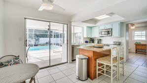 A kitchen with a central island, a dining area with wicker chairs, and a view of a swimming pool through glass doors.