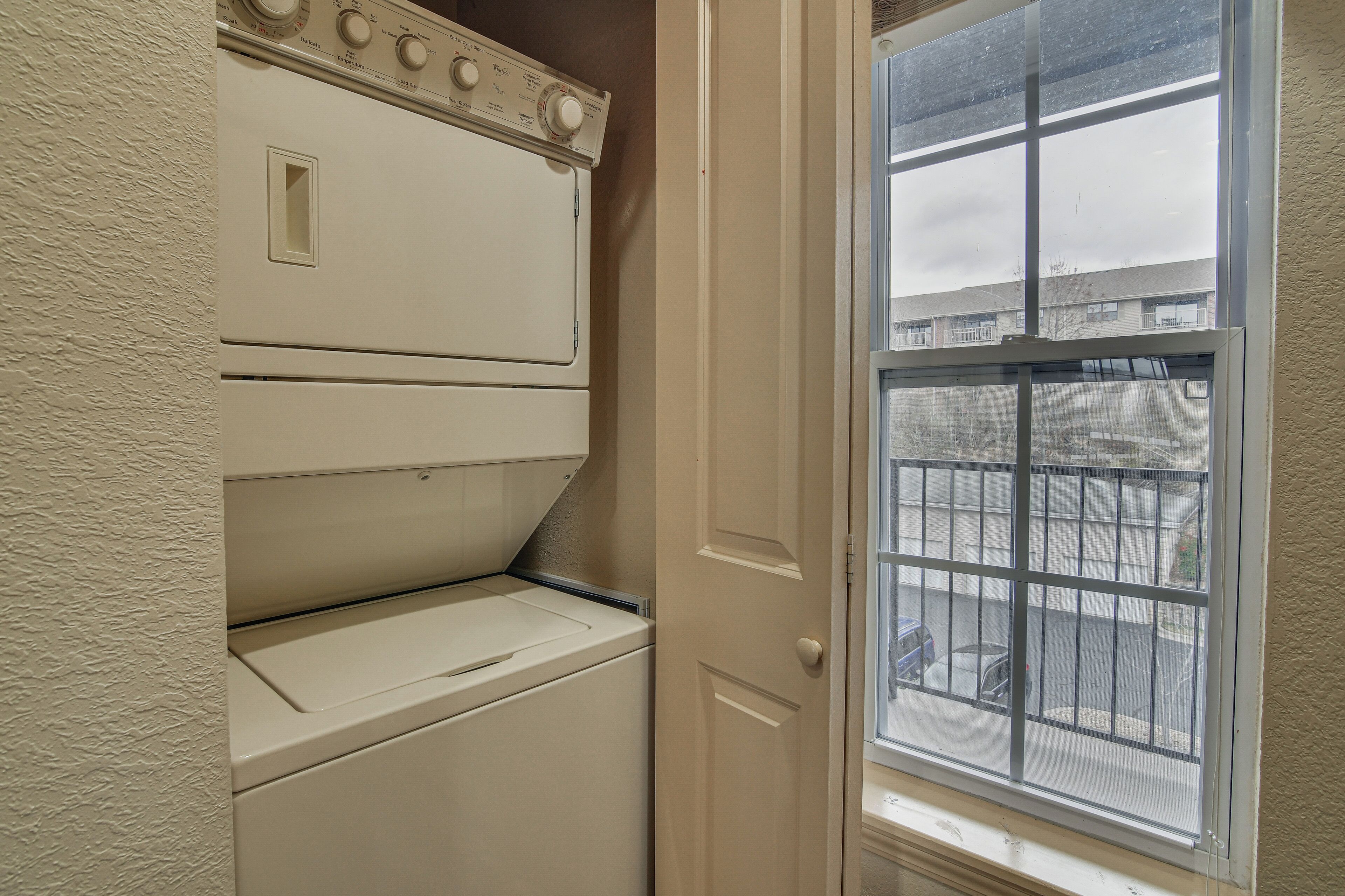 Une buanderie équipée d’une machine à laver et d’un sèche-linge superposés, et une porte donnant sur un balcon avec vue sur un quartier résidentiel.